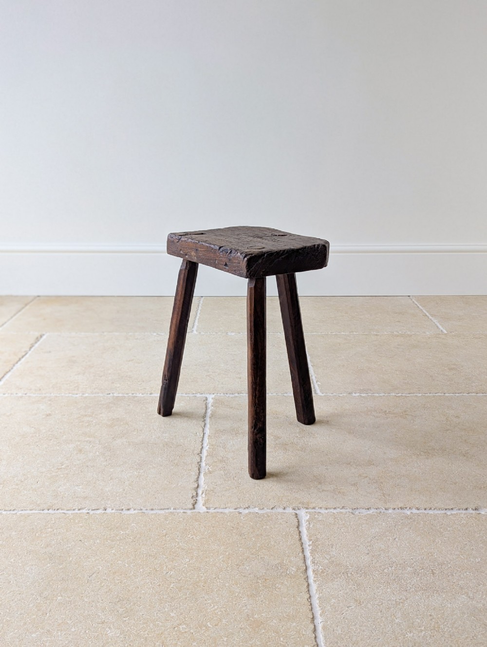 A dark oak Victorian three-legged stool featuring a thick rectangular top and weathered splayed legs.