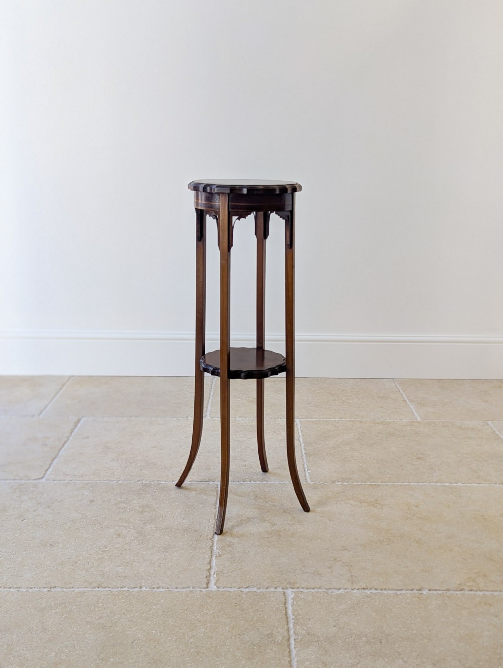 A tall, slender Edwardian mahogany torchere featuring a circular scalloped top, geometric line inlay, flared legs, and a small lower tier.