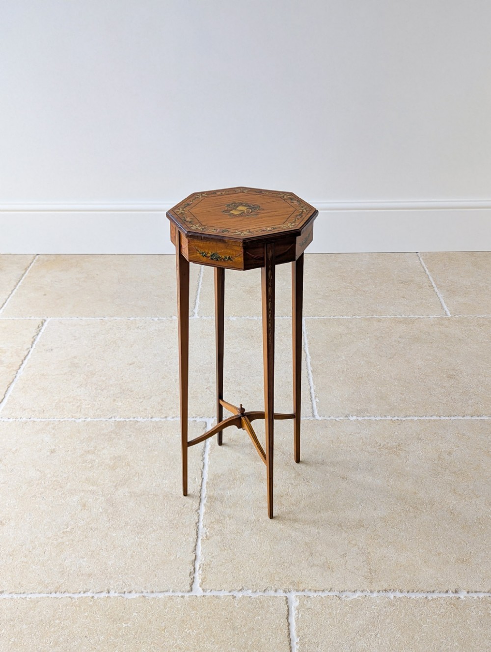 An Edwardian octagonal satinwood candle stand featuring painted floral borders, four slender tapered legs, and an X-form stretcher with a central finial.