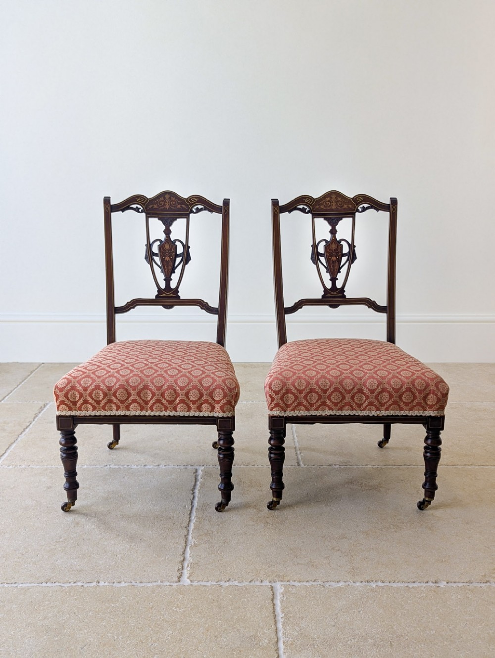 A pair of Edwardian mahogany salon chairs featuring urn marquetry splats, turned front legs on brass castors, and patterned upholstery.