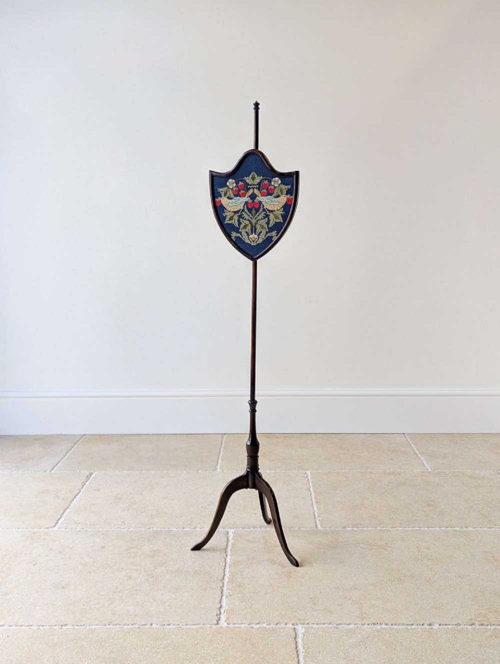 An antique George III mahogany pole screen featuring a tripod base, turned column, and a shield-shaped needlework panel depicting birds and strawberries.