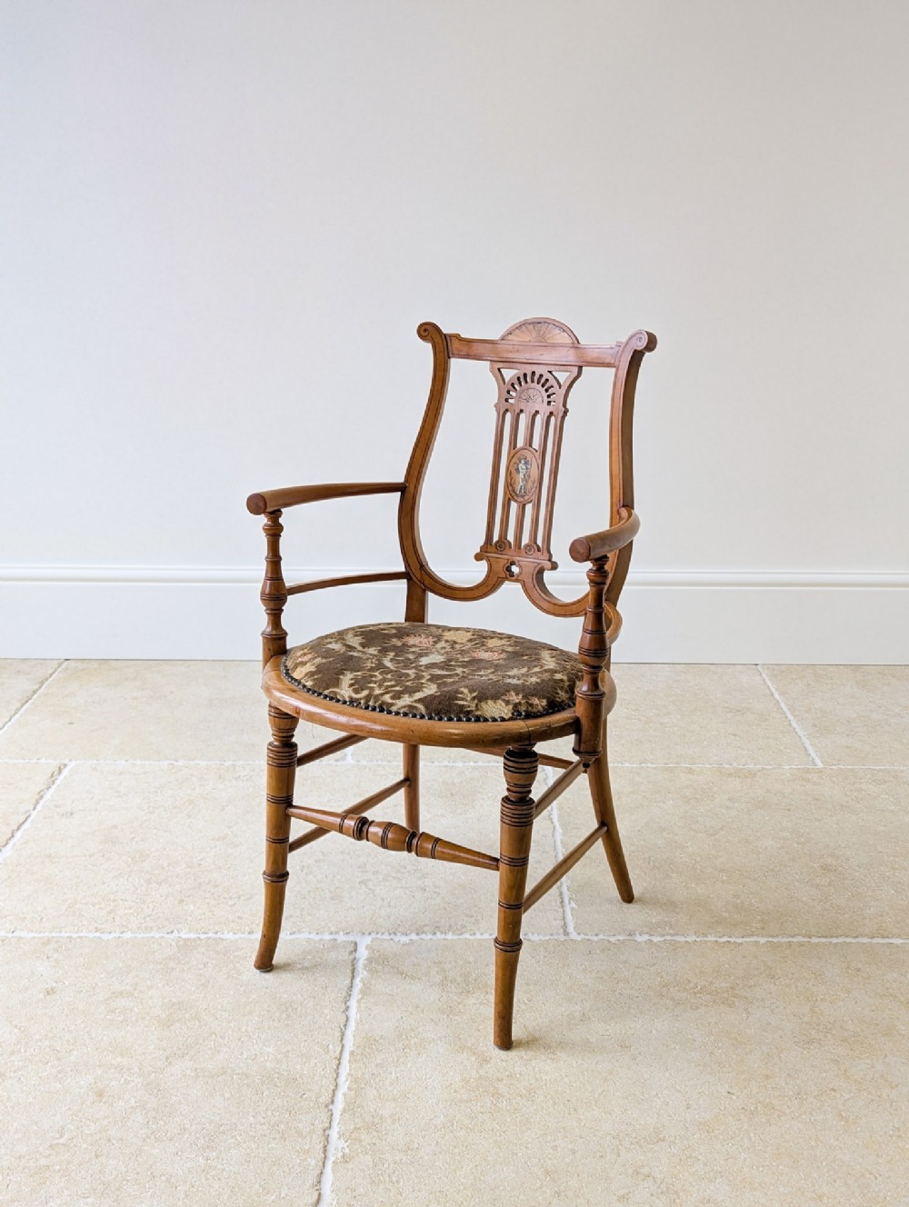 An antique satinwood armchair featuring a carved lyre-shaped backrest with marquetry inlay, turned arms and legs, and a floral upholstered seat.