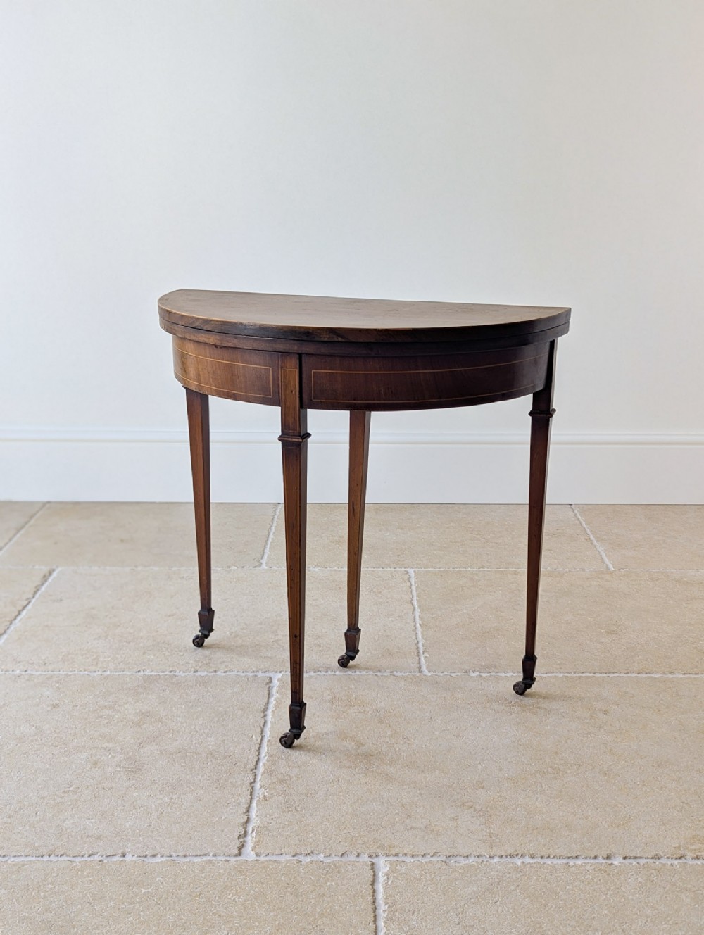 An Edwardian mahogany demilune card table featuring inlaid stringing detail, square tapered legs, and small brass castors.