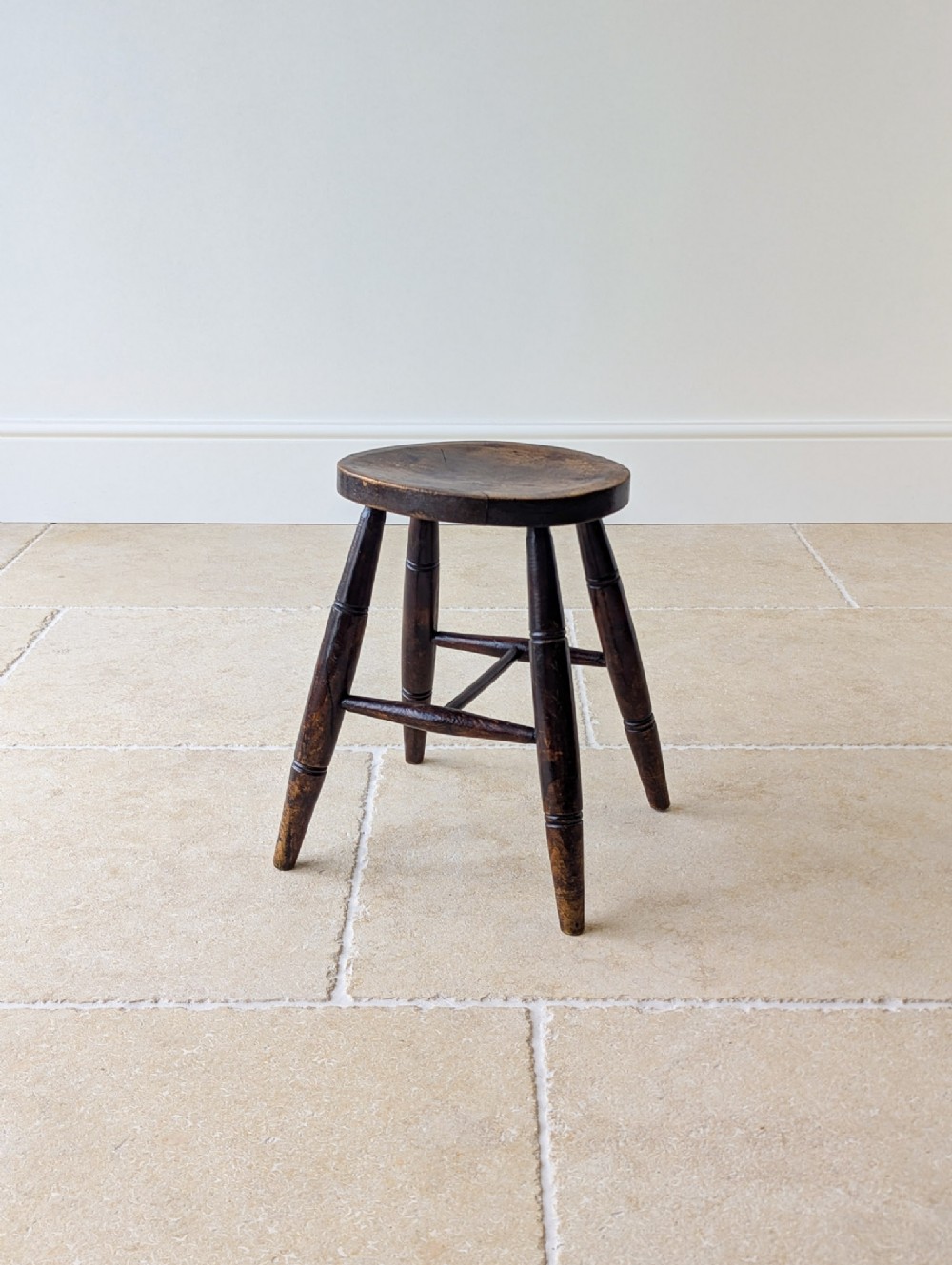 An antique dark oak country stool with a thick circular seat, turned splayed legs, and cross stretchers.
