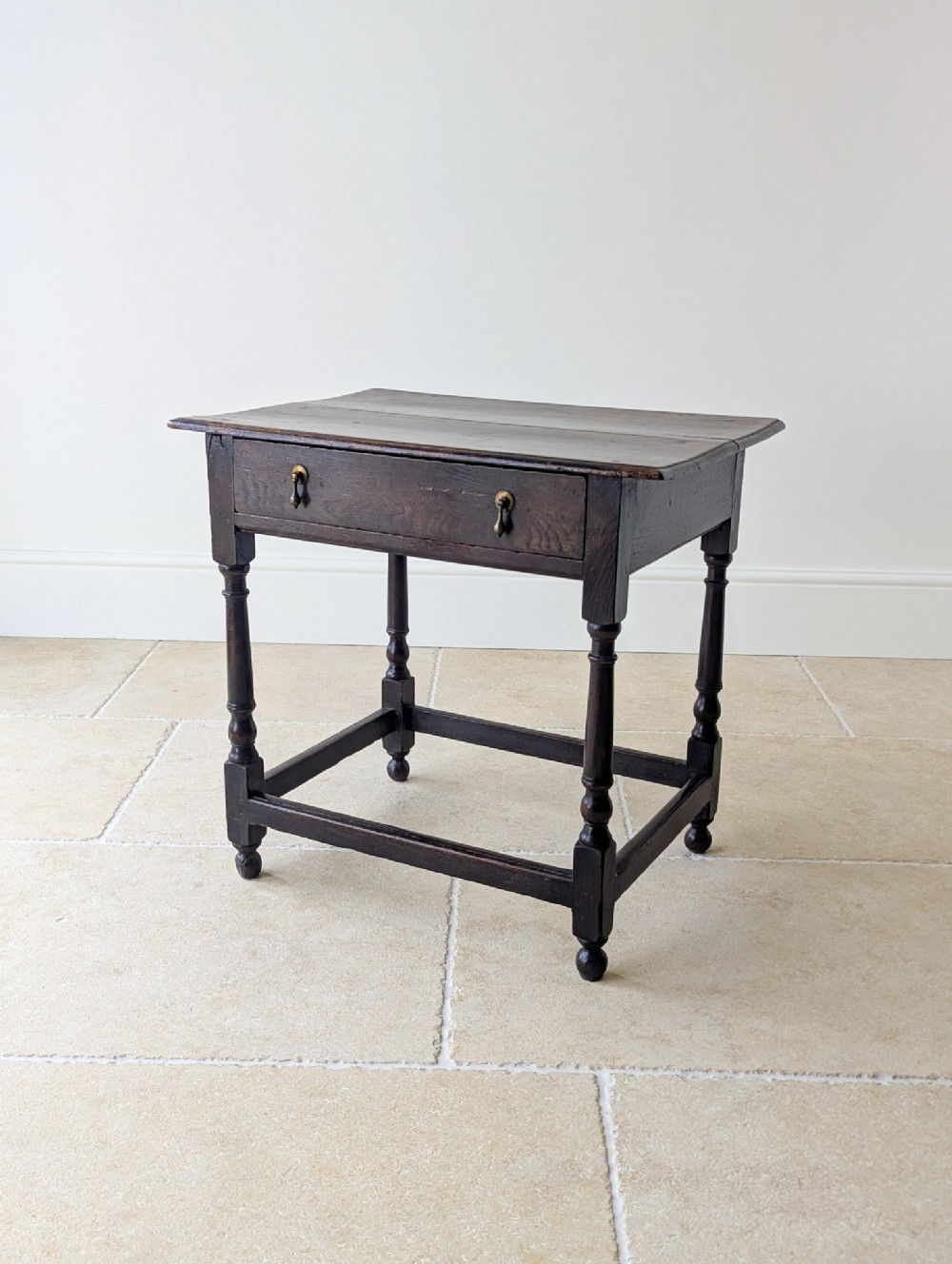 A late 17th-century dark oak side table featuring a single front drawer with brass teardrop handles, turned legs, and a lower rectangular box stretcher.