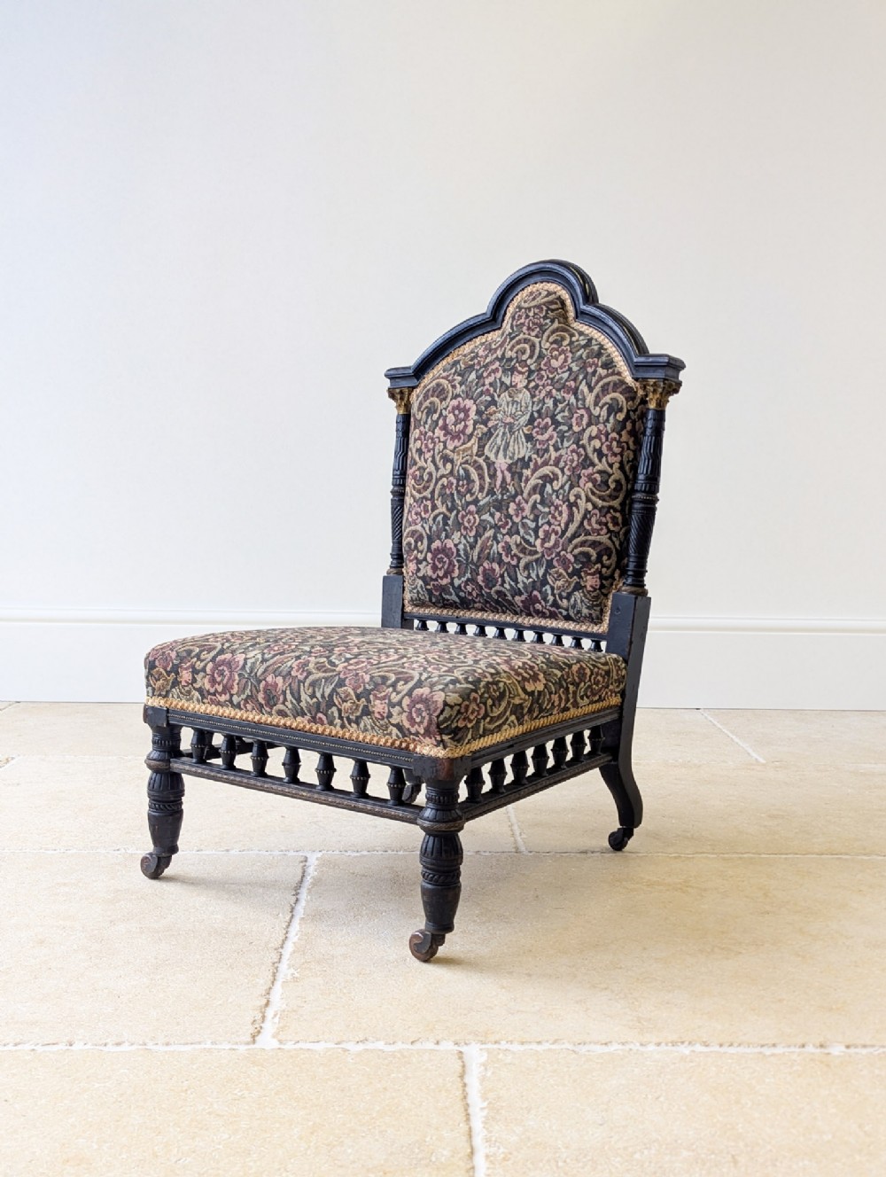 A low-seated Victorian Aesthetic Movement chair featuring ebonised turned wood detailing, a gallery of spindles, floral tapestry upholstery, and front brass castors.
