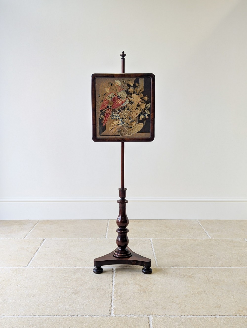 A William IV rosewood pole screen featuring a square needlepoint panel displaying a parrot and flowers, set on a turned column with a triform base.
