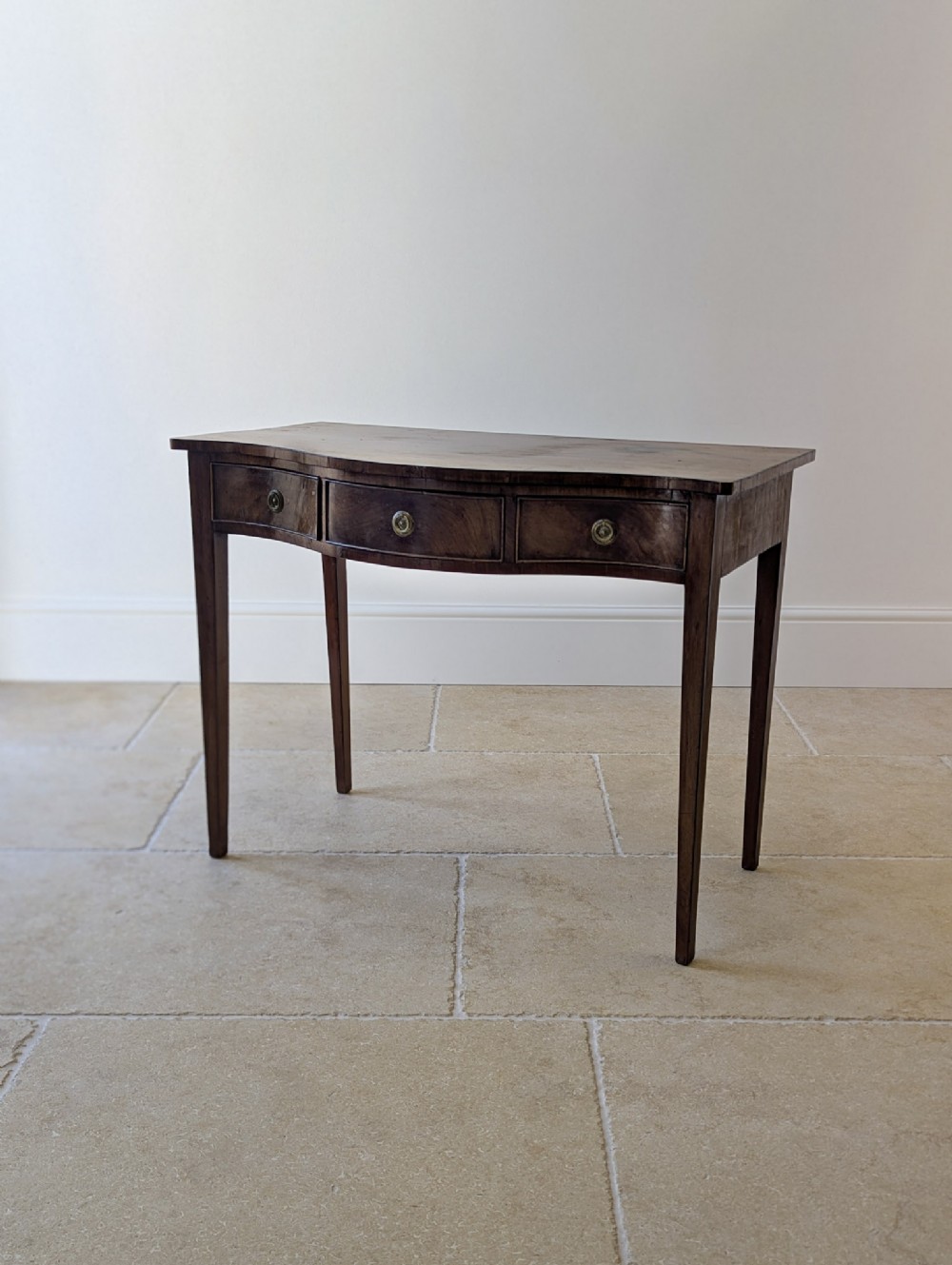 A Georgian mahogany side table featuring a serpentine front profile, three drawer fronts with circular brass handles, and four tapered legs.