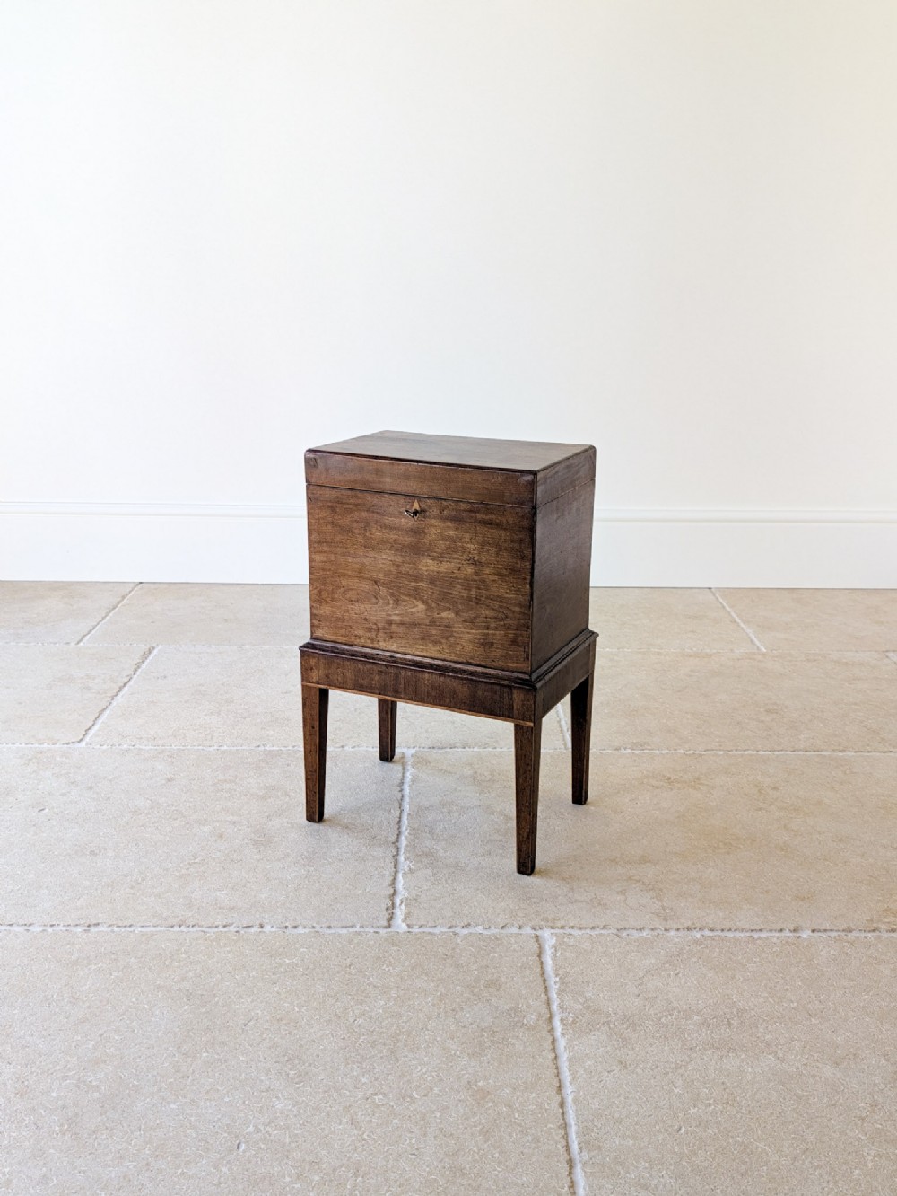 An antique George III dark mahogany cellaret chest featuring a hinged top, inlaid escutcheon, and square tapered legs on a stone tile floor.