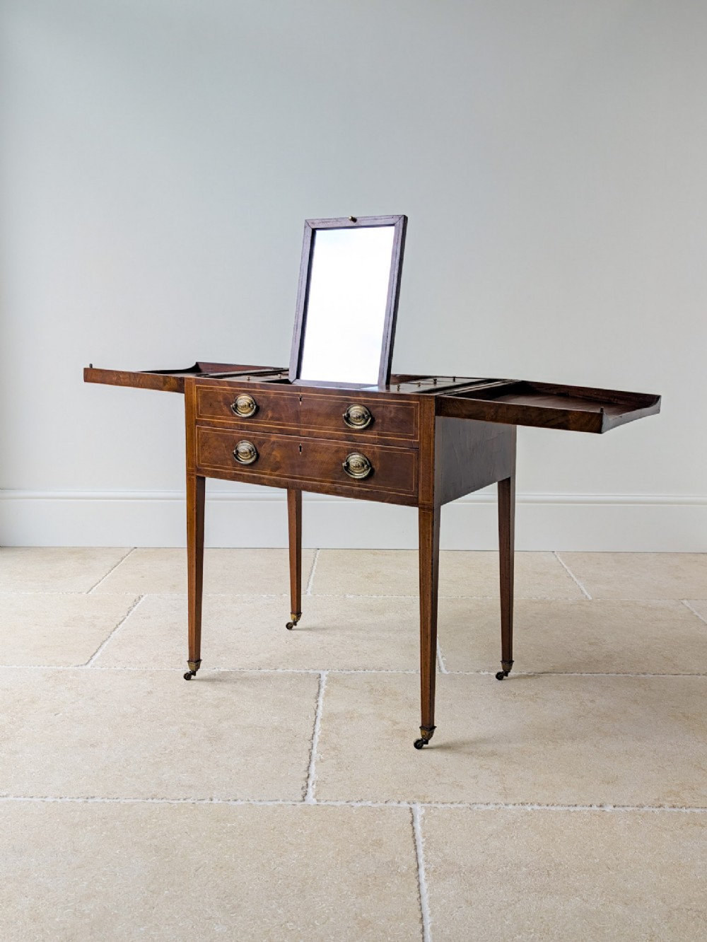 An antique Georgian mahogany folding dressing table with fold-out side tops, a central pop-up mirror, brass handles, and square tapered legs on castors.