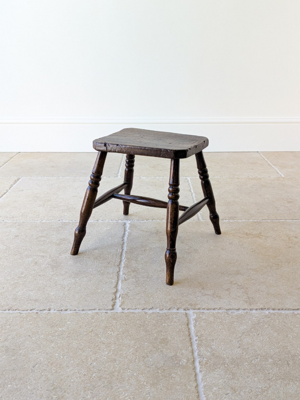 A dark brown antique Victorian country stool made of elm and beech with turned wooden legs and a solid rectangular top.