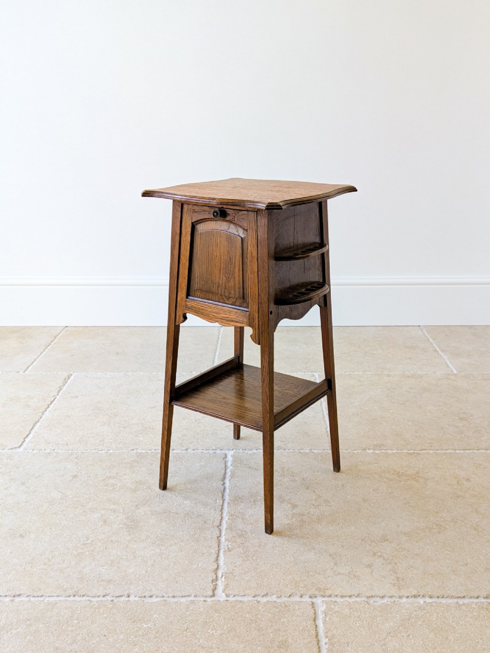 An antique Arts and Crafts solid oak occasional table featuring an overhanging top, a central cupboard door, side racks, and a lower tier shelf on slender square legs.