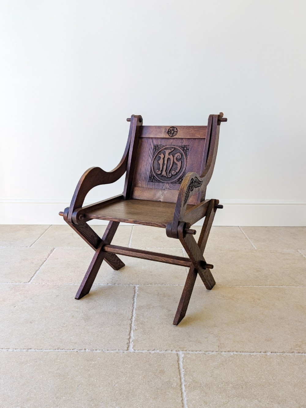 An Edwardian dark oak Glastonbury armchair featuring an X-frame base, wooden peg construction, and a carved IHS monogram back panel.