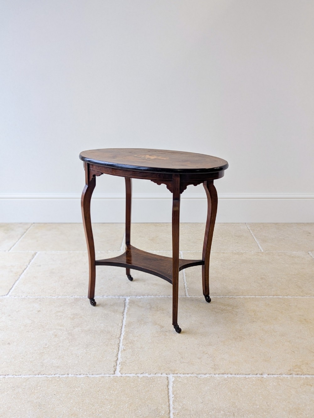 An Edwardian oval rosewood occasional table featuring slender cabriole legs, a lower undertier, marquetry inlay on top, and small brass castors.