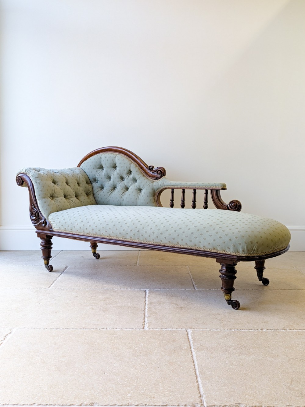 A Victorian mahogany chaise longue featuring sage green button-tufted upholstery, carved scroll details, turned spindle arm, and legs supported by brass castors.