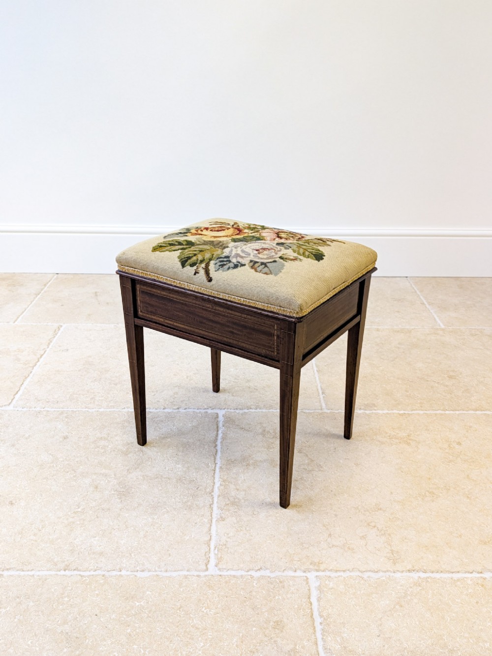 An Edwardian dark mahogany piano stool featuring tapered square legs, boxwood stringing inlay, and a floral needlepoint upholstered seat.