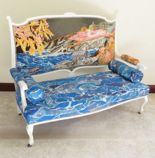 fig 1. An Art Nouveau settee featuring a white painted wooden frame, a backrest upholstered with a detailed pictorial coastal landscape scene, and a seat upholstered in a blue marbled wave pattern.