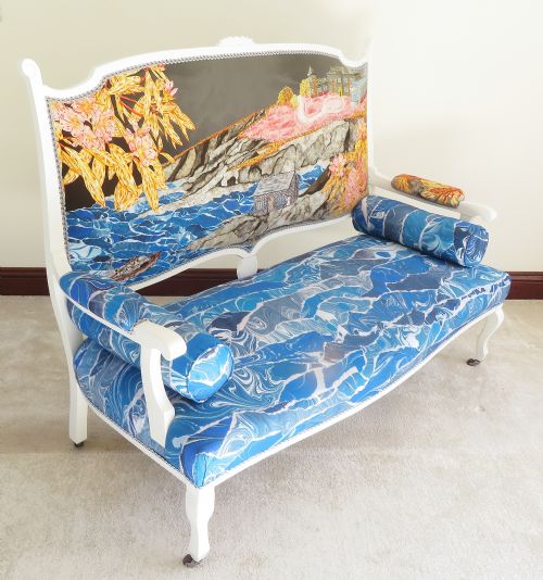 fig 2. An Art Nouveau settee featuring a white painted wooden frame, a backrest upholstered with a detailed pictorial coastal landscape scene, and a seat upholstered in a blue marbled wave pattern.