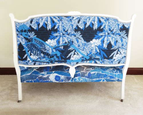 fig 3. An Art Nouveau settee featuring a white painted wooden frame, a backrest upholstered with a detailed pictorial coastal landscape scene, and a seat upholstered in a blue marbled wave pattern.