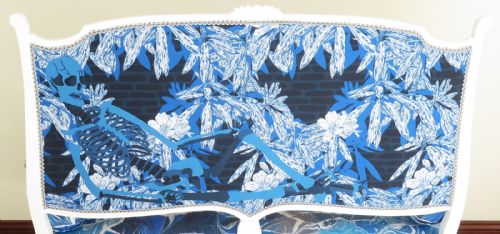 fig 5. An Art Nouveau settee featuring a white painted wooden frame, a backrest upholstered with a detailed pictorial coastal landscape scene, and a seat upholstered in a blue marbled wave pattern.
