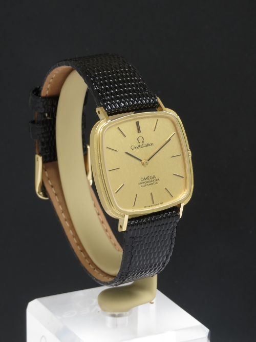 A 1968 Omega Constellation Deluxe wristwatch featuring a cushion-shaped 18ct gold case, brushed champagne dial, baton indices, and a black textured leather strap. View VI