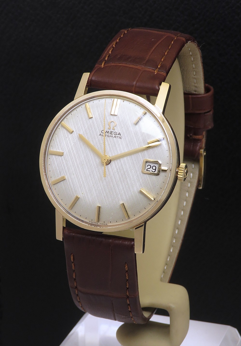 A 1965 solid 9ct gold Omega automatic wristwatch featuring a textured silver dial, gold baton hour markers, and a brown leather strap.