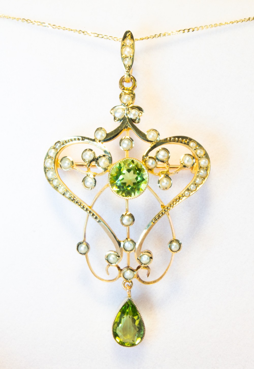 An openwork gold Art Nouveau pendant featuring curved scrollwork set with seed pearls, a central circular green peridot, and a hanging pear-cut peridot drop.