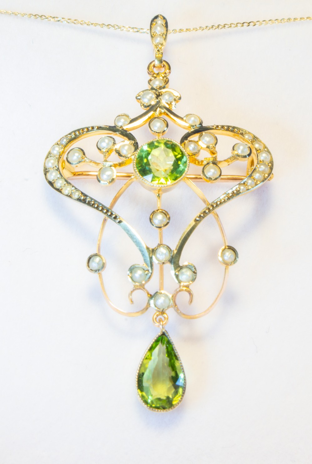 An openwork gold Art Nouveau pendant featuring curved scrollwork set with seed pearls, a central circular green peridot, and a hanging pear-cut peridot drop. View II