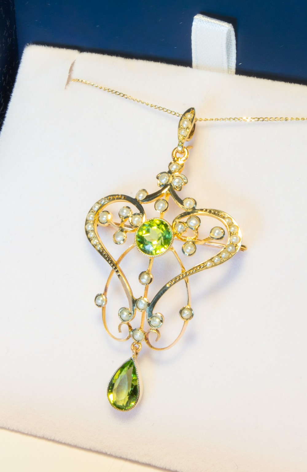An openwork gold Art Nouveau pendant featuring curved scrollwork set with seed pearls, a central circular green peridot, and a hanging pear-cut peridot drop. View III