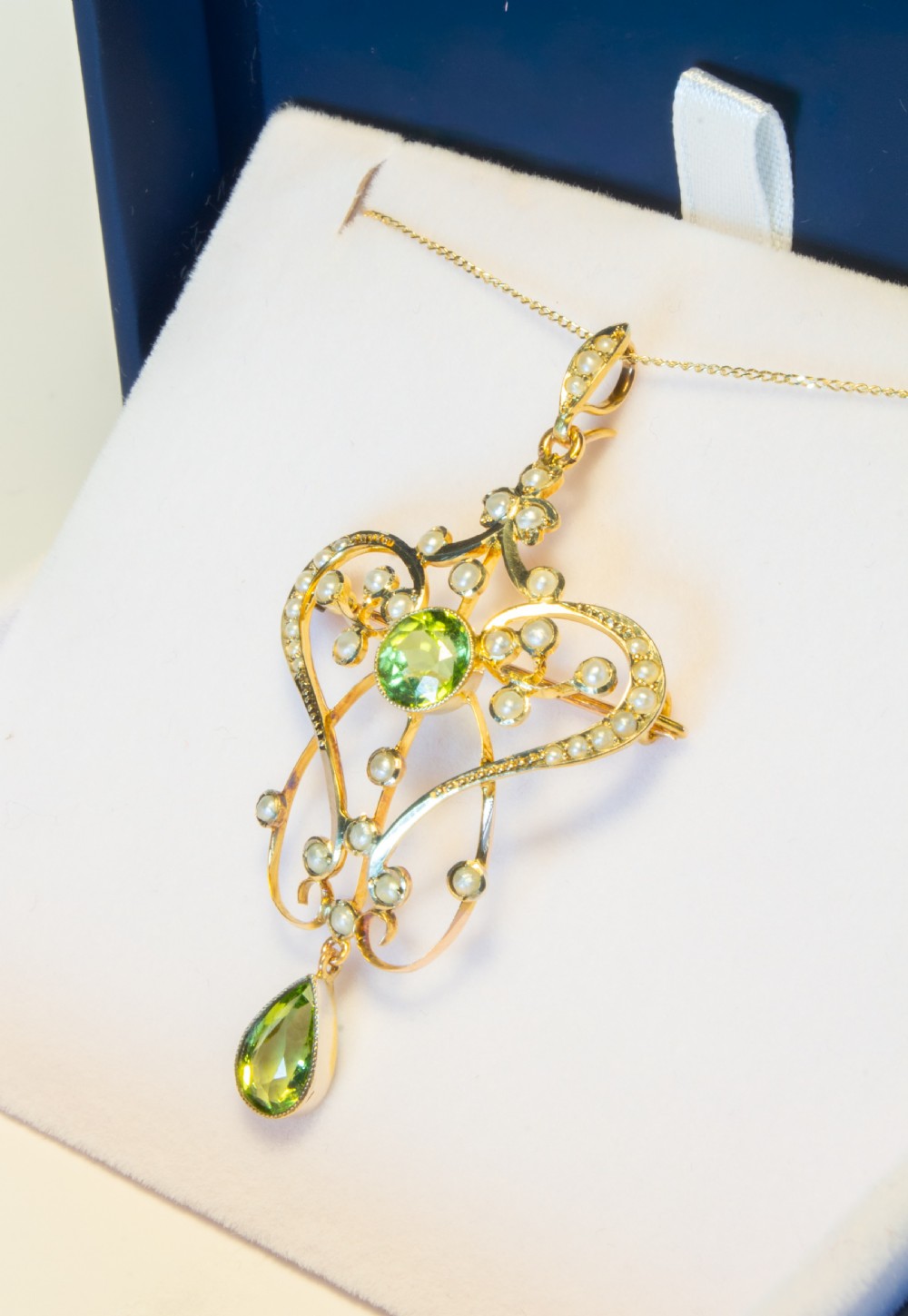 An openwork gold Art Nouveau pendant featuring curved scrollwork set with seed pearls, a central circular green peridot, and a hanging pear-cut peridot drop. View IV