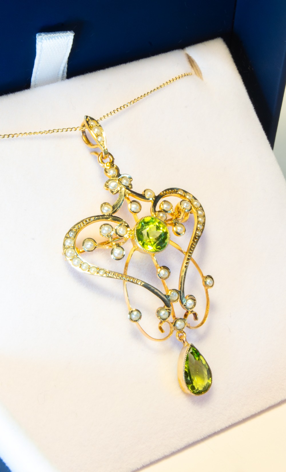 An openwork gold Art Nouveau pendant featuring curved scrollwork set with seed pearls, a central circular green peridot, and a hanging pear-cut peridot drop. View VI
