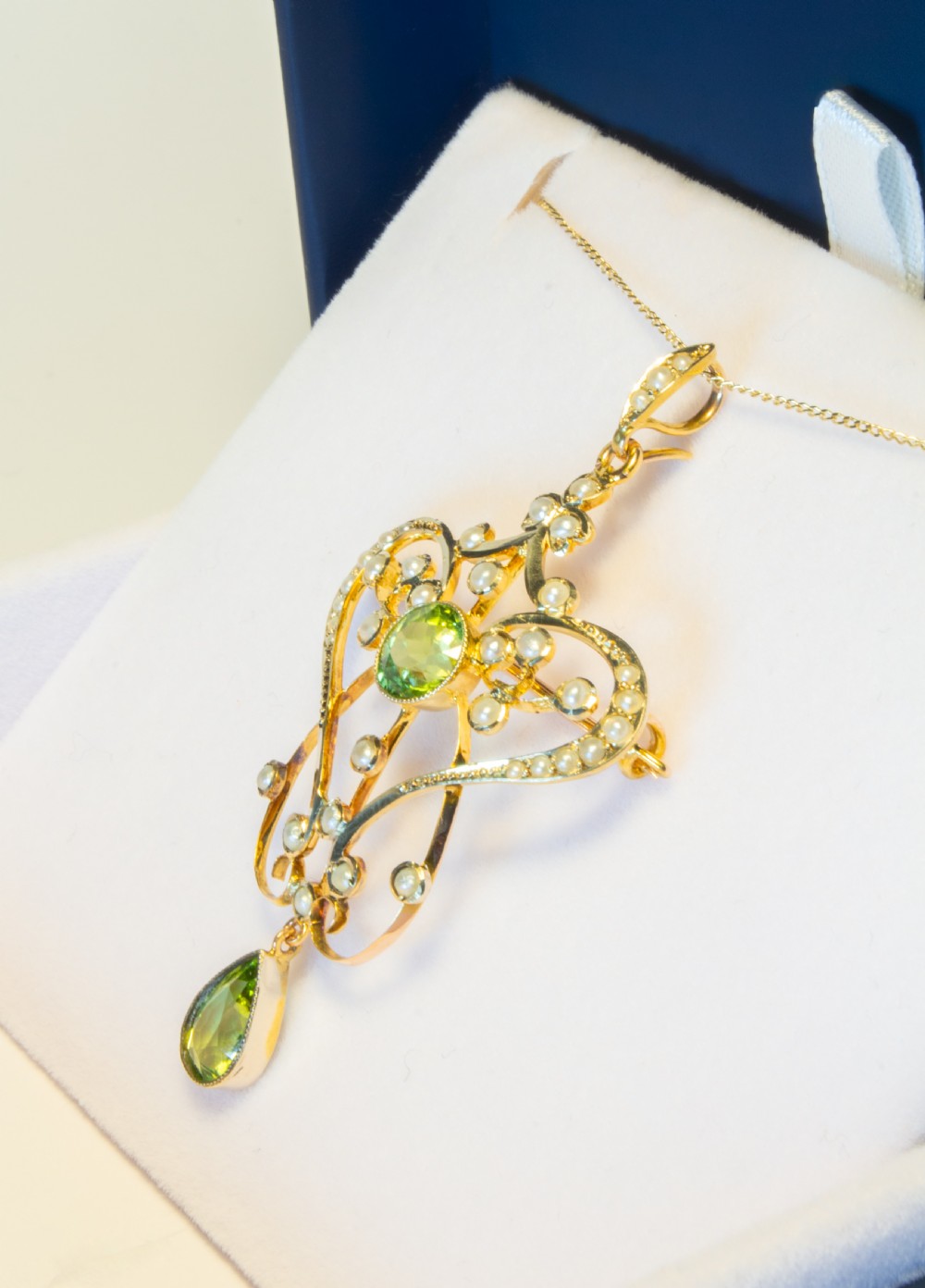 An openwork gold Art Nouveau pendant featuring curved scrollwork set with seed pearls, a central circular green peridot, and a hanging pear-cut peridot drop. View VII