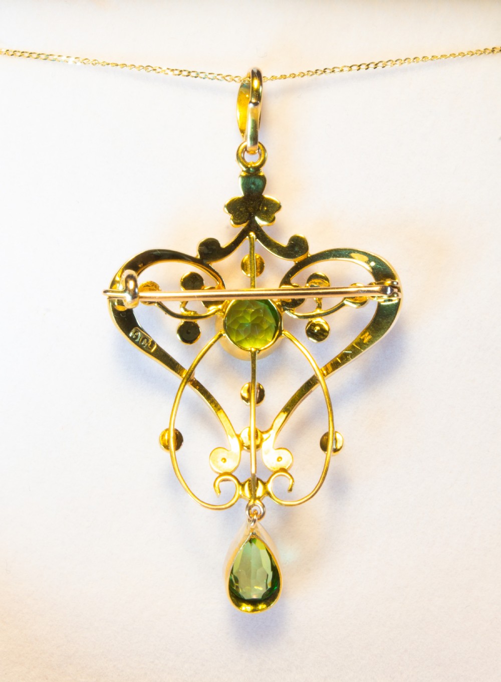 An openwork gold Art Nouveau pendant featuring curved scrollwork set with seed pearls, a central circular green peridot, and a hanging pear-cut peridot drop. View VIII