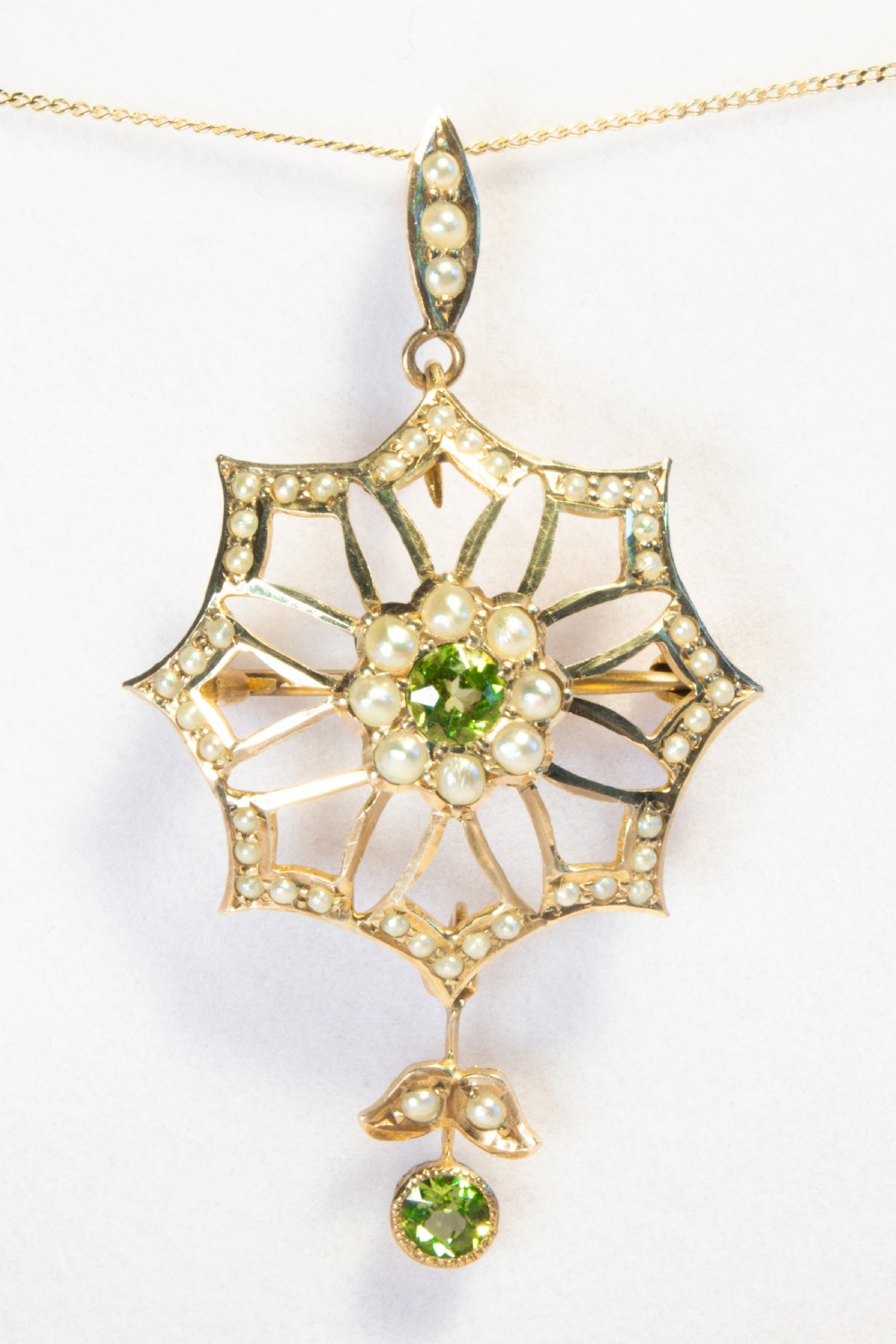 An openwork gold starburst pendant brooch set with seed pearls and a central round peridot, featuring a suspended drop peridot.