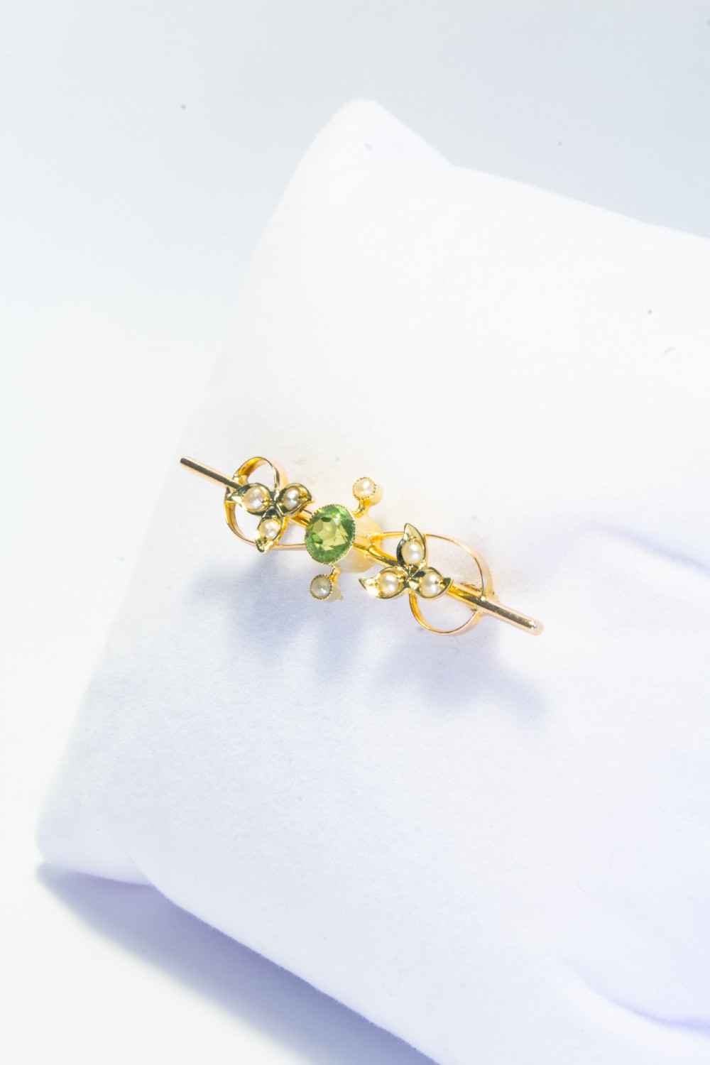An early 20th-century 9ct gold bar brooch featuring a central round facetted green peridot flanked by foliate leaf motifs set with seed pearls. View V