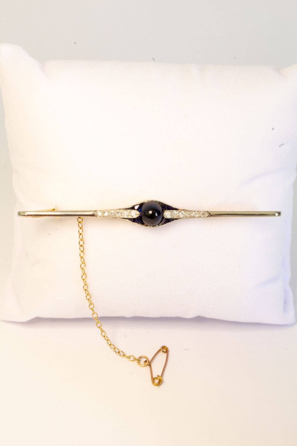 An 18ct gold and platinum bar brooch featuring a central dark cabochon sapphire flanked by small diamonds and calibre-cut sapphires with an attached safety chain. View II