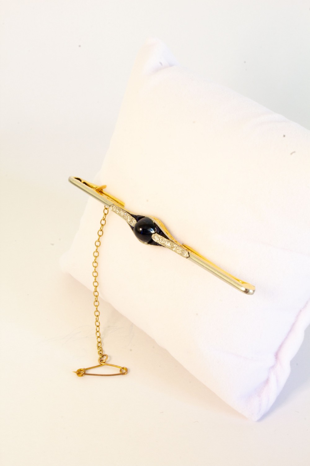An 18ct gold and platinum bar brooch featuring a central dark cabochon sapphire flanked by small diamonds and calibre-cut sapphires with an attached safety chain. View IV