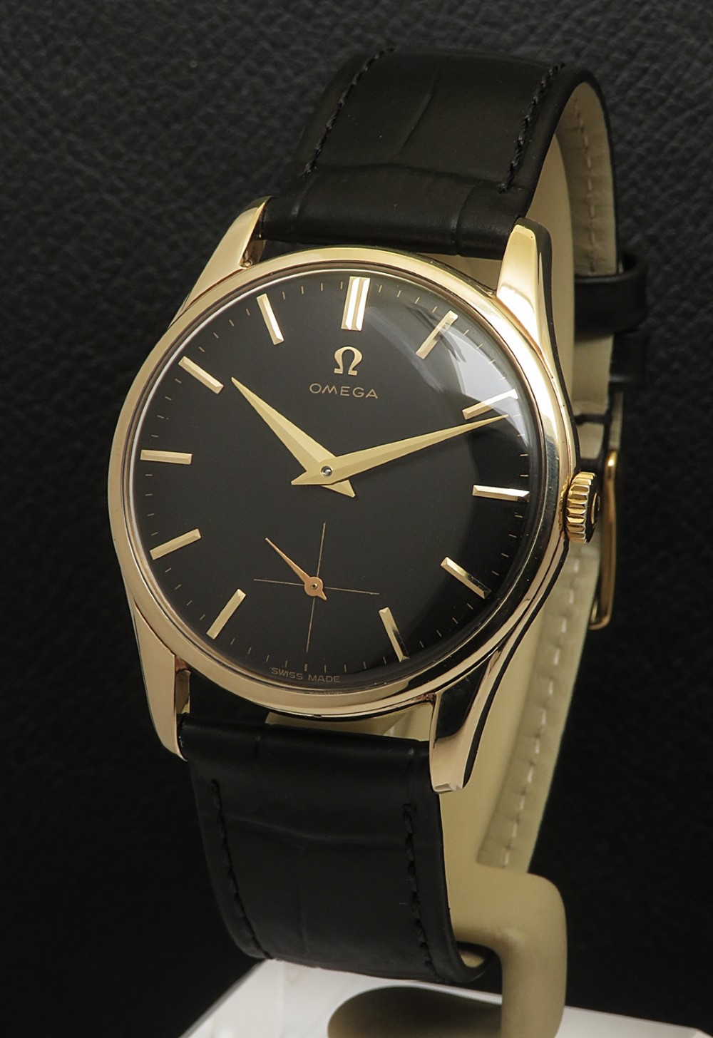 A vintage Omega wristwatch featuring a solid gold circular case, a black dial with gold baton hour markers, gold dauphine hands, a small seconds sub-dial, and a black leather strap.