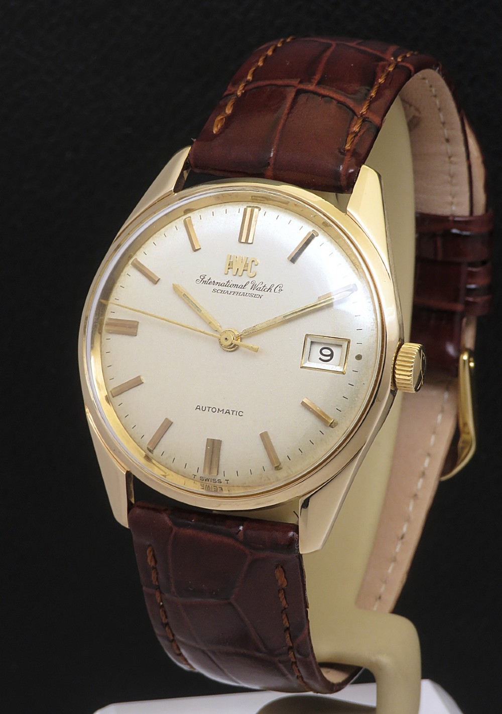 A 1969 solid 9ct gold IWC automatic wristwatch featuring a silvered dial, gold baton markers, date window, and brown leather strap.