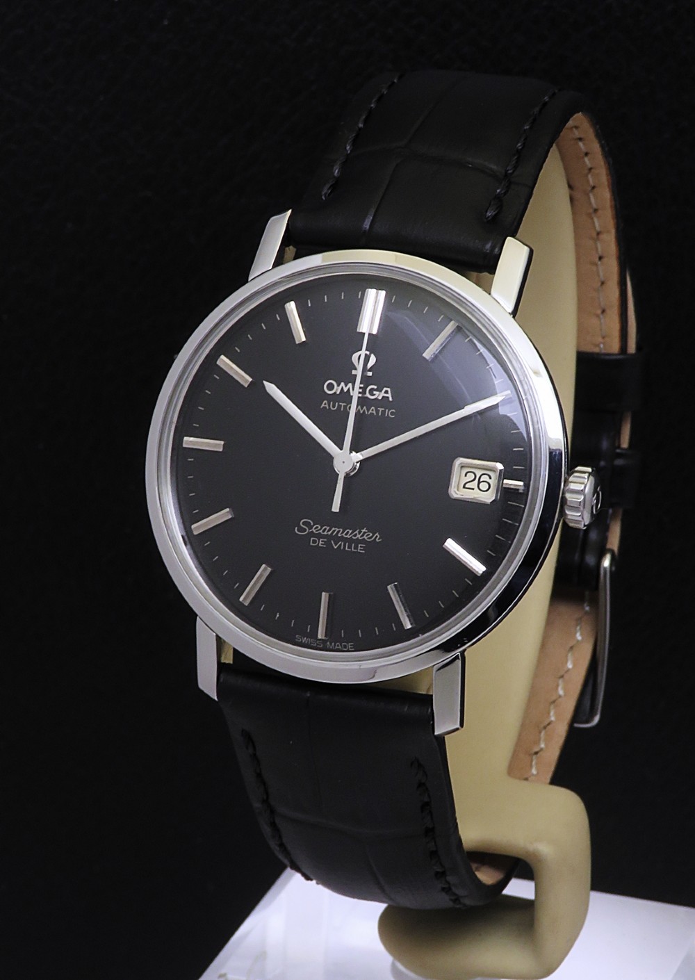 A 1965 Omega Seamaster DeVille vintage wristwatch featuring a round stainless steel case, a black dial with silver baton hour markers, a date window at three oclock, and a black leather strap.