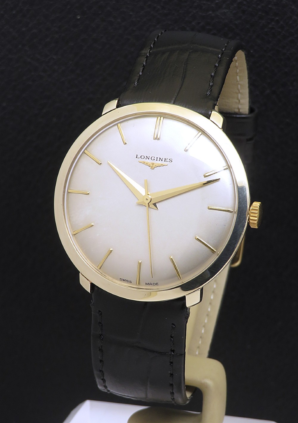 A 1960 vintage Longines wristwatch featuring a round solid gold case, silver dial with gold markers, and a black leather strap.