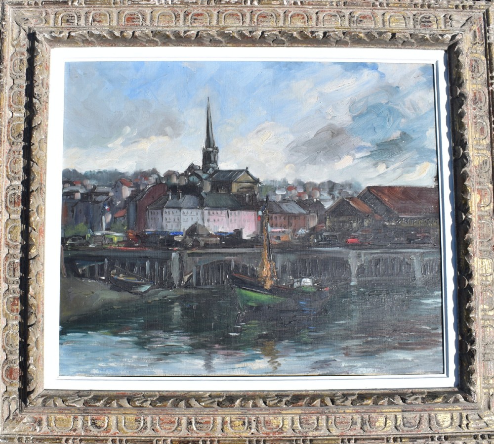 An impressionist oil painting depicting boats anchored in a French harbour against a backdrop of town buildings and a spire, set within an ornate carved frame.