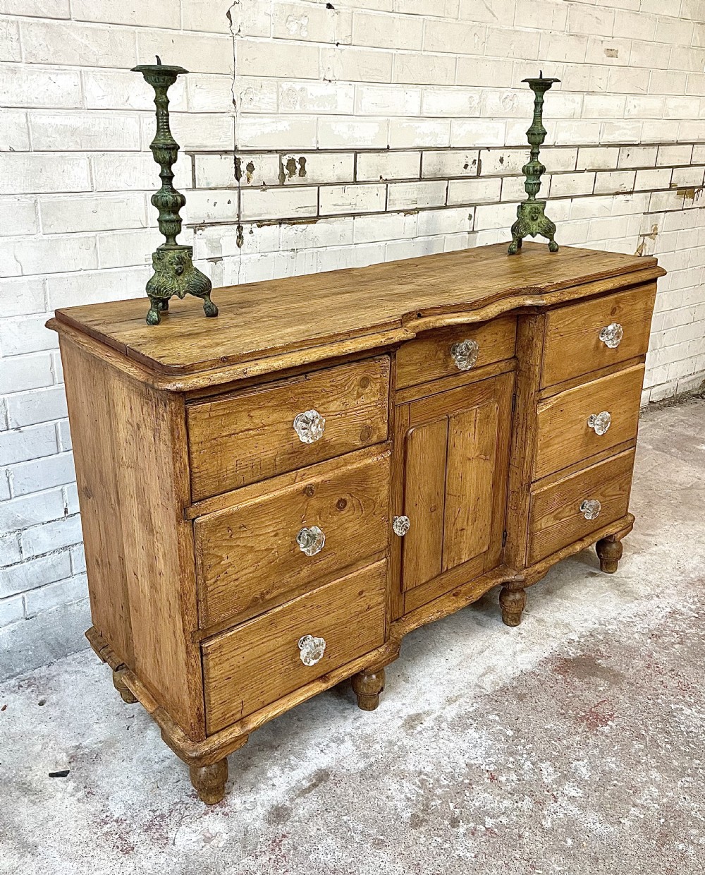 A warm brown pine sideboard featuring a scalloped top edge, six side drawers, a central drawer, a panelled cupboard door, and clear glass knobs.