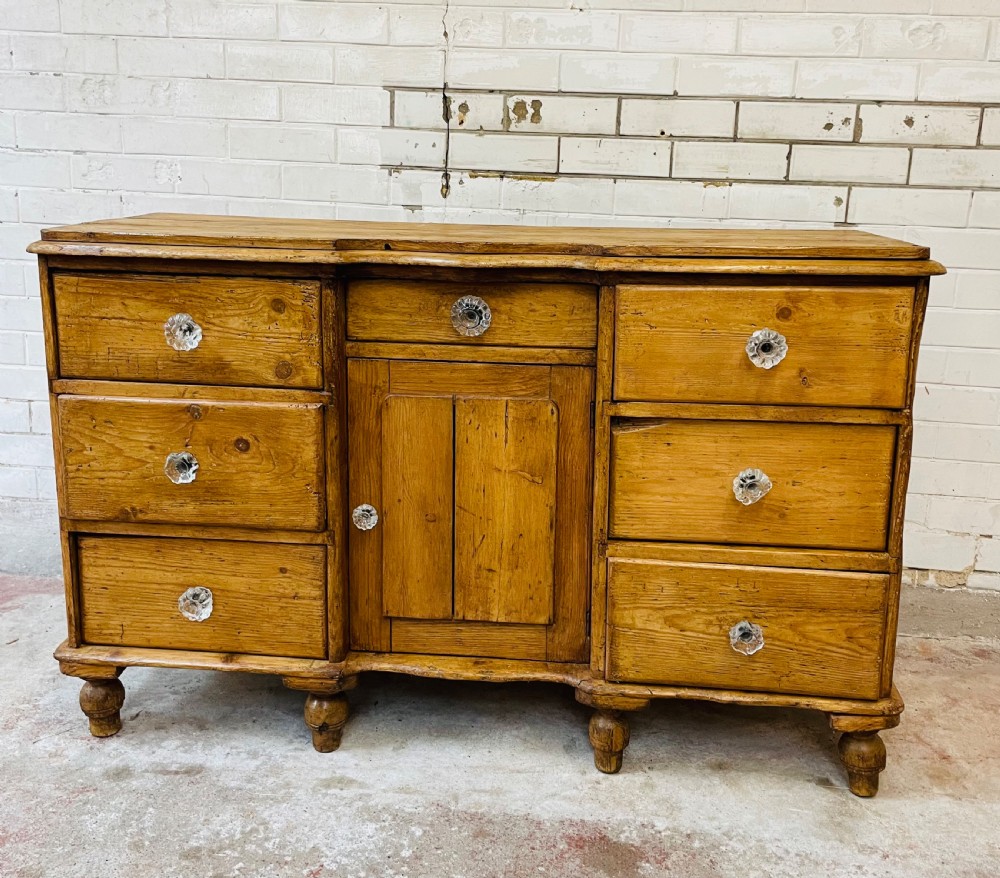 A warm brown pine sideboard featuring a scalloped top edge, six side drawers, a central drawer, a panelled cupboard door, and clear glass knobs. View III