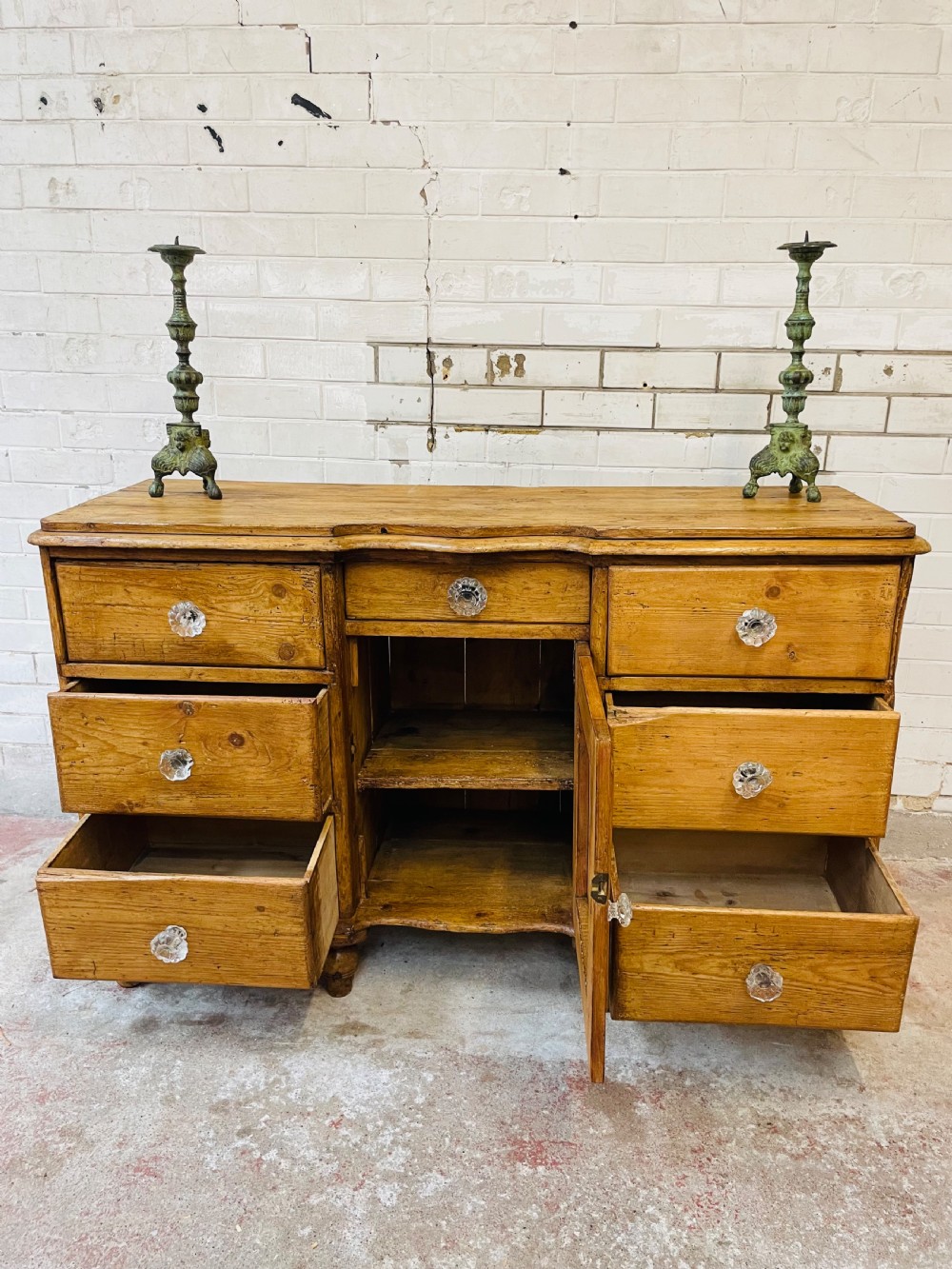 A warm brown pine sideboard featuring a scalloped top edge, six side drawers, a central drawer, a panelled cupboard door, and clear glass knobs. View IV
