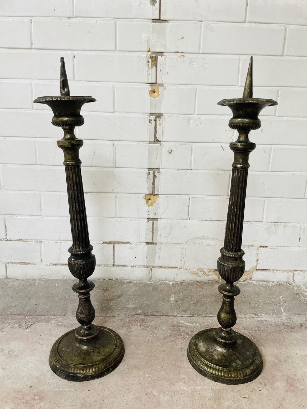 A pair of tall aged brass pricket candlesticks featuring fluted central columns, flared drip trays, top candle spikes, and circular patterned bases. View II