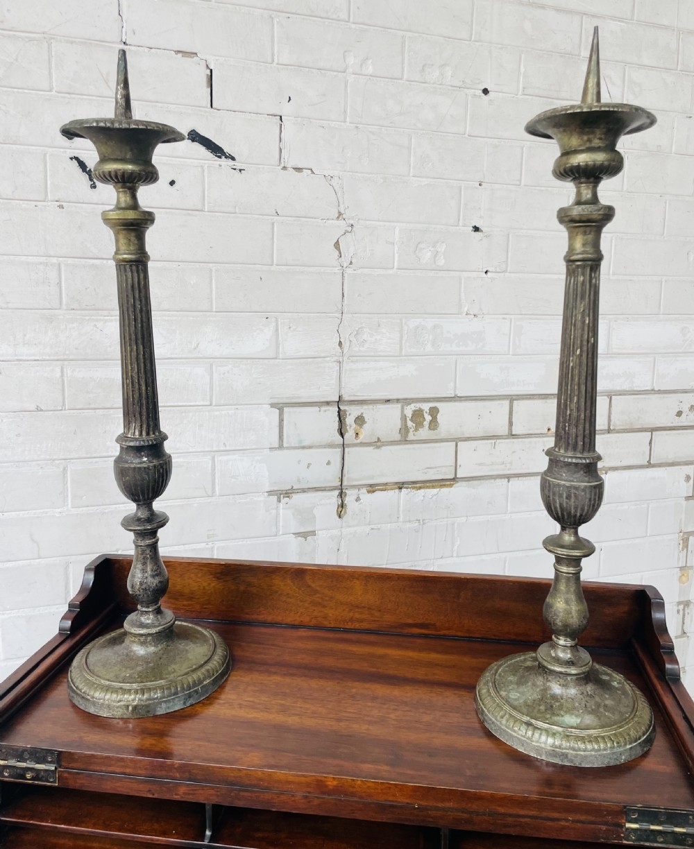 A pair of tall aged brass pricket candlesticks featuring fluted central columns, flared drip trays, top candle spikes, and circular patterned bases. View VI