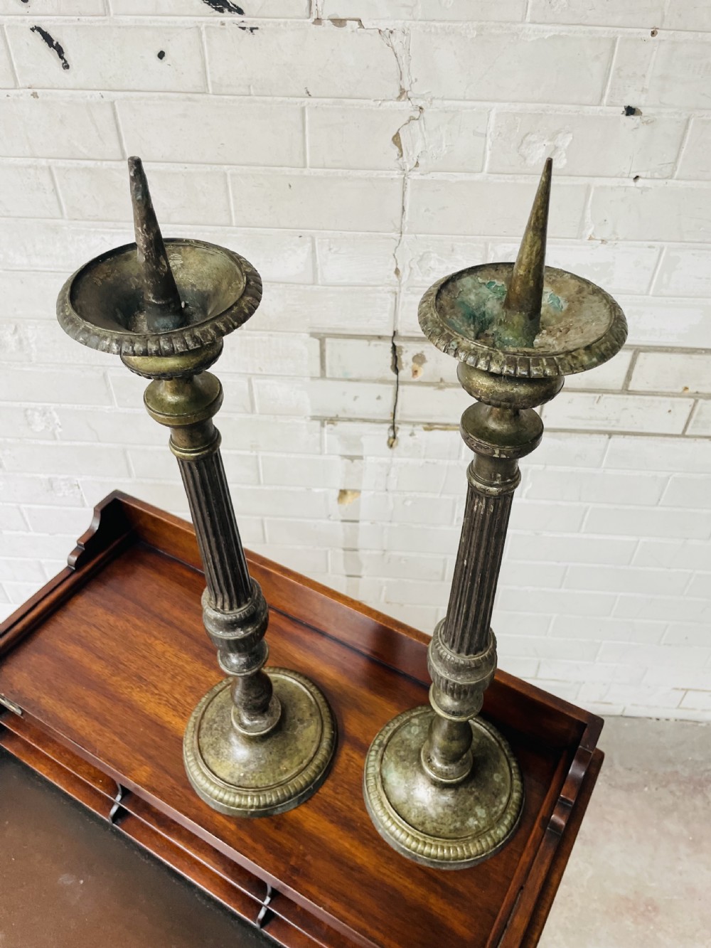 A pair of tall aged brass pricket candlesticks featuring fluted central columns, flared drip trays, top candle spikes, and circular patterned bases. View VII