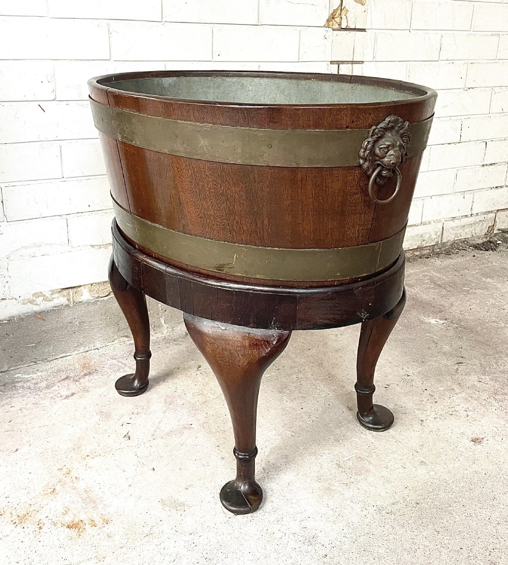 An oval brass-bound mahogany wine cooler on a stand with cabriole legs, pad feet, and lion mask handles.