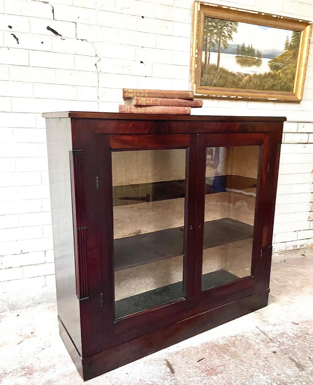 A low dark mahogany glazed bookcase featuring two glass panel doors, fluted side pilasters, and an integrated plinth base.
